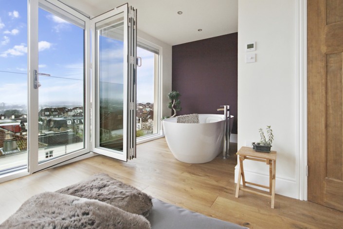 Bi-fold doors open on a glass juliet balcony showing an attractive suburban view on a clear day. A luxury bath in an l-shaped bedroom looks out over the city or the stars.