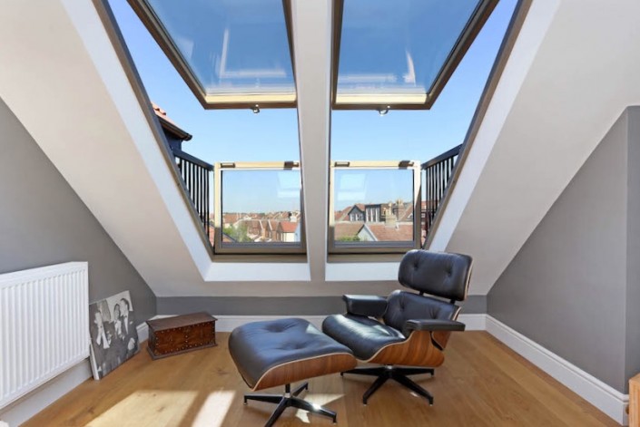 A stylish designer leather and wood Eames chair with matching foot stool waits under double, open, Velux Cabrio Balcony windows. Inside there is a medium toned wood floor and white and grey walls. Outside the sky is blue.
