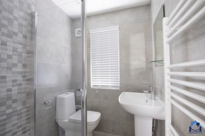 Landscape orientation photograph of a small en-suite shower room. The shower cubicle is barely visible to the left, beyond it is a modern loo and to the right a full sized pedestal basin, both in white. The oblong window in the rear wall features venetian blinds. The whol room is tiled in matt grey with some large mosaic-style tiles that blend in.