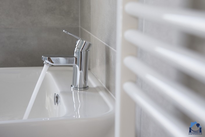 Landscape orientation image of bathroom basin mono tap running water just off centre to the left, facing left, with blurred white heated towel rail leading into the picture from the right.