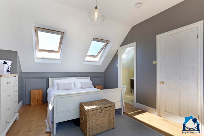 View of a double bedroom in a loft conversion, showing double bed with two Velux roof windows above. The room is decorated white for the ceilings and sloped walls, grey walls and mid tone natural wood floors.