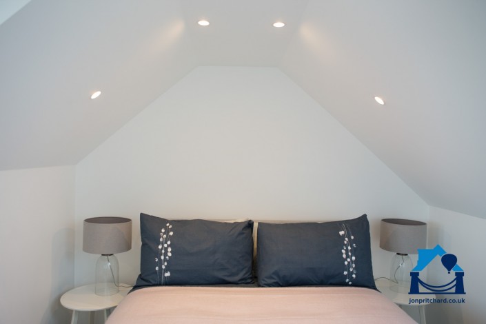 Feature of top end of double bed with pillows in dark blue, under the eaves of a loft conversion, with spotlights