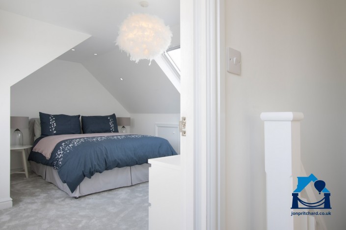 Image showing a loft conversion master bedroom, taken through the door from the landing. A bright room carpeted in light grey, with navy bedding and VELUX windows visible